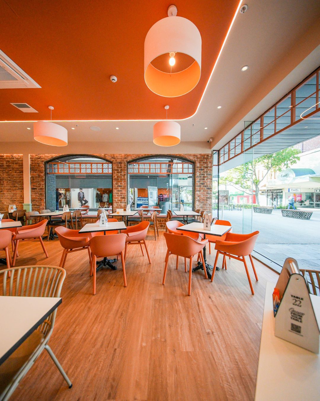 A completed café fit-out — orange tub chairs and tables under drum pendants, exposed brick and an open shopfront onto the street.